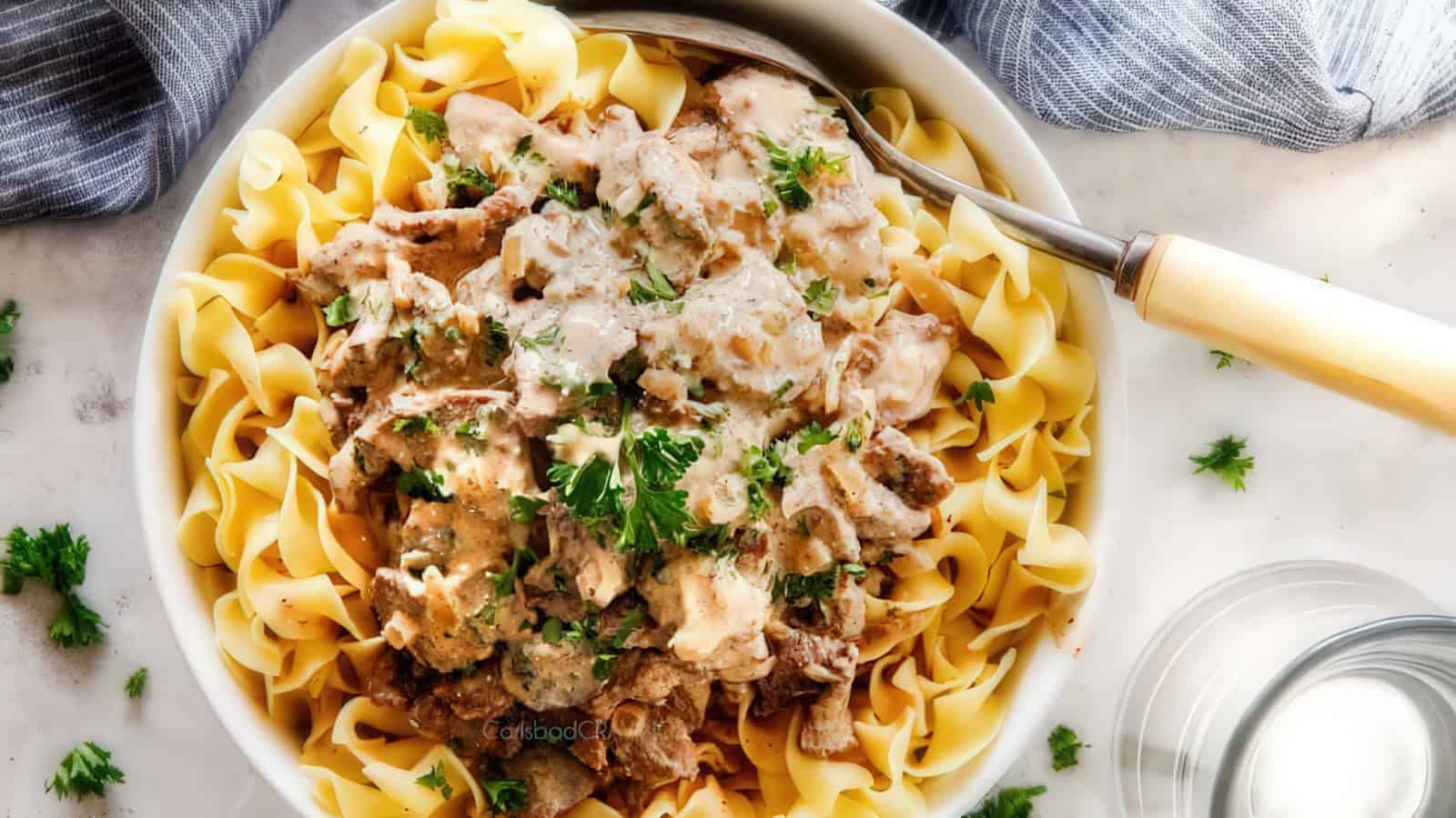 A bowl of pasta topped with creamy mushroom and meat sauce, garnished with fresh parsley. A spoon rests on the side, and a glass of water nearby. A striped cloth is in the background.