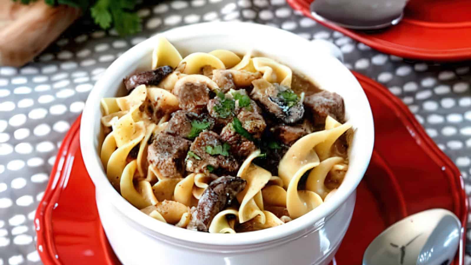 A bowl of beef stroganoff with tender beef, egg noodles, and garnished with fresh parsley. It's placed on a red plate, with a silver spoon nearby. The background has a patterned cloth.