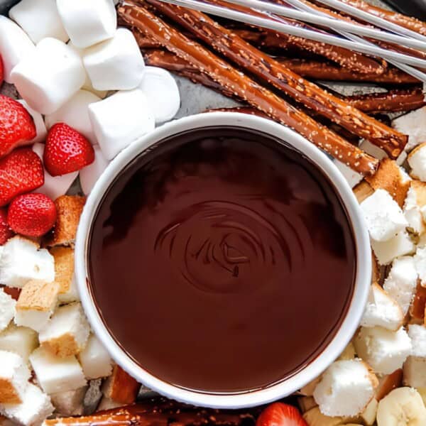 A platter with a bowl of chocolate fondue in the center, surrounded by marshmallows, strawberries, cubes of cake, pretzel rods, and banana slices, all arranged for dipping. Skewers are laid beside the bowl on a round tray.