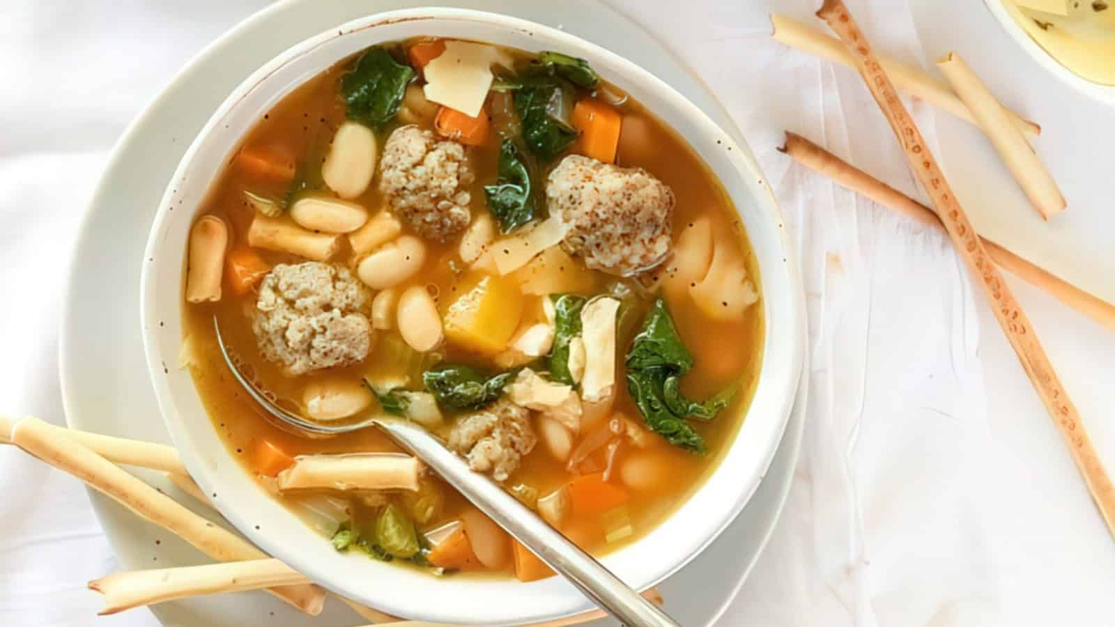 A bowl of soup with meatballs, white beans, diced carrots, leafy greens, and pieces of cheese, accompanied by a few breadsticks on the side. A spoon rests in the bowl, all set on a light-colored tablecloth.