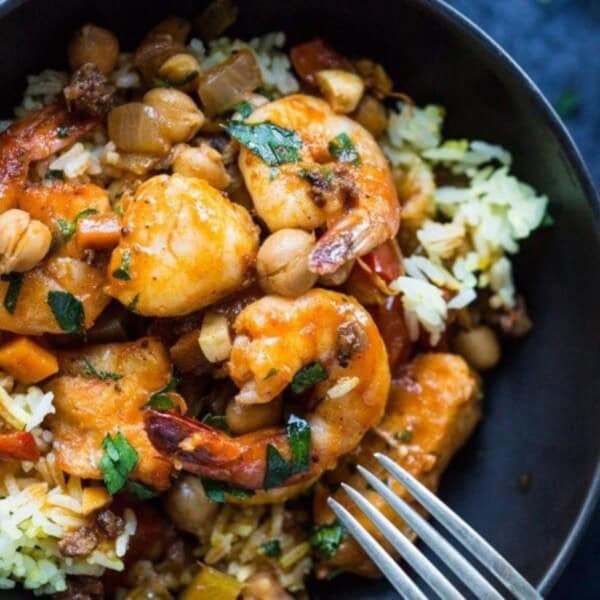 A bowl of flavorful shrimp curry with rice, chickpeas, and vegetables. The dish is garnished with fresh herbs and served in a black bowl, with a fork resting on the side. The vibrant colors of the ingredients make the dish visually appealing.