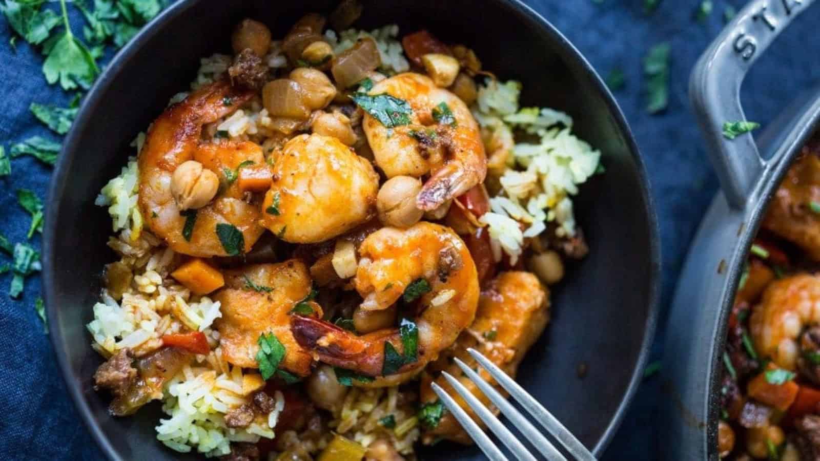 A bowl of flavorful shrimp curry with rice, chickpeas, and vegetables. The dish is garnished with fresh herbs and served in a black bowl, with a fork resting on the side. The vibrant colors of the ingredients make the dish visually appealing.