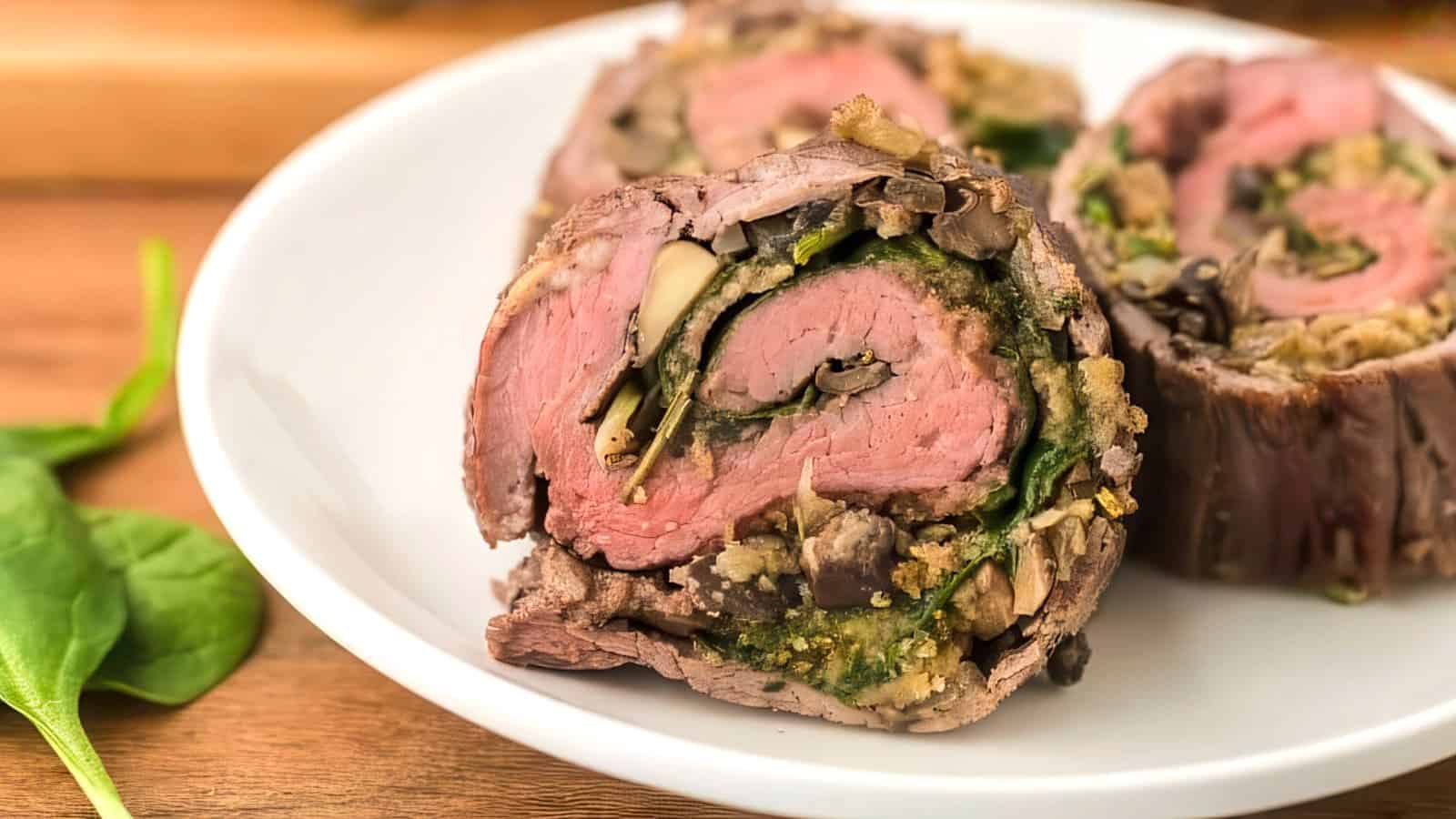 Sliced beef roulade on a white plate, filled with spinach, mushrooms, and other vegetables, showcasing a cross-section of the rolled meat and colorful filling. Some fresh spinach leaves are visible on the wooden surface beside the plate.