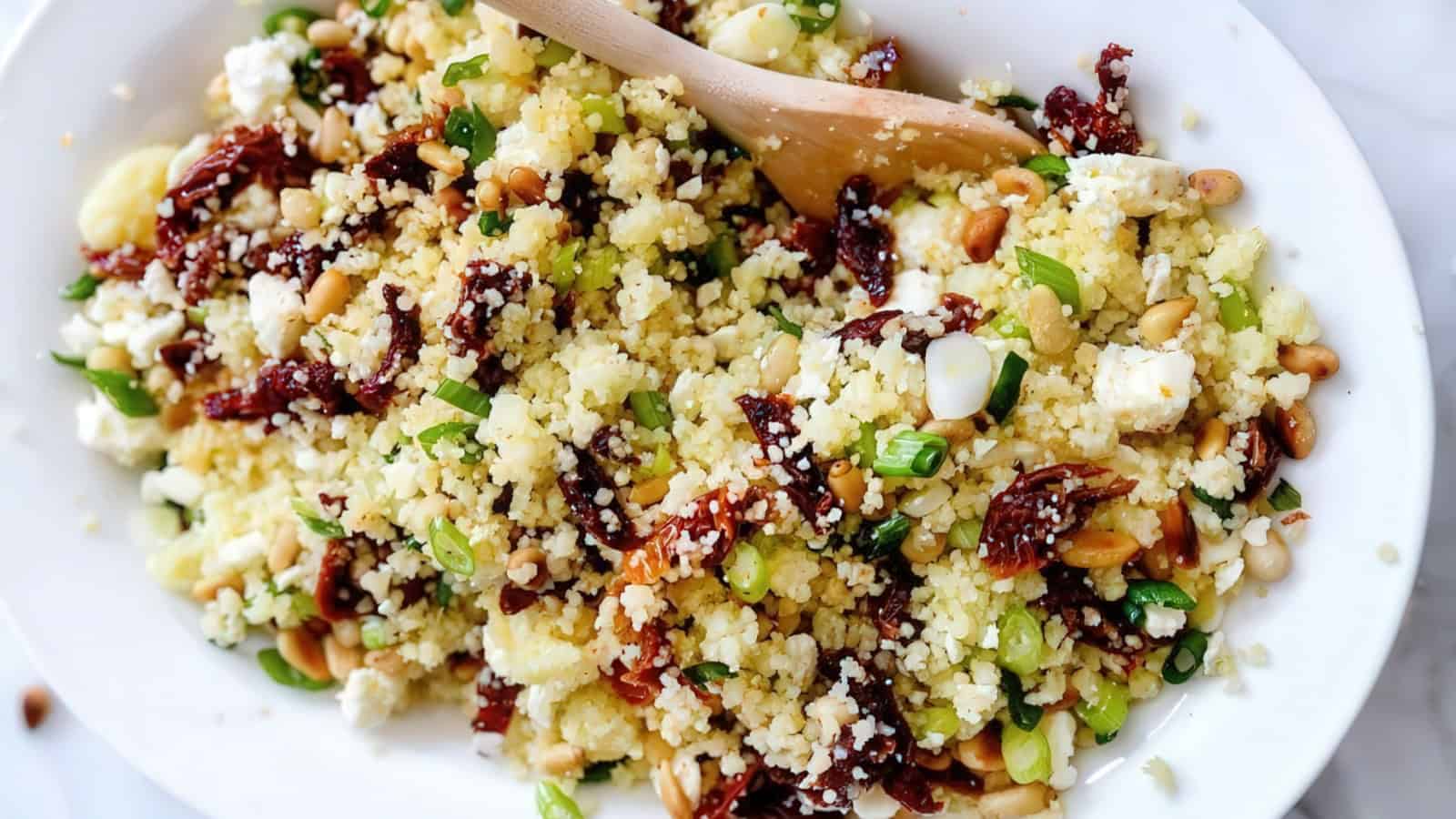 A plate of couscous salad with sun-dried tomatoes, feta cheese, pine nuts, and chopped green onions, mixed with a wooden spoon. The bowl sits on a white surface.
