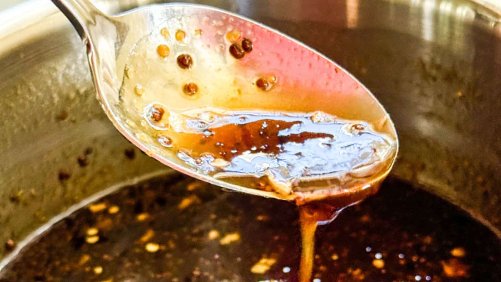A close-up of a spoon dripping with thick, dark, homemade balsamic glaze, held over a pot. The glaze is shiny and smooth, with small bits of seasoning visible.