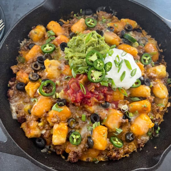 A cast iron skillet containing baked tater tots topped with ground beef, melted cheese, black olives, salsa, sour cream, guacamole, sliced jalapeños, and green onions.