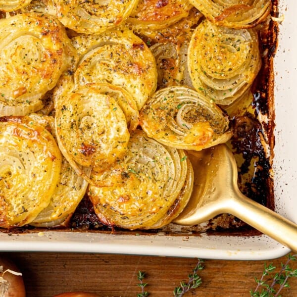 Sliced onions in a white rectangular dish with browned edges, topped with herbs and cheese, gold serving spoon and onion peels nearby.