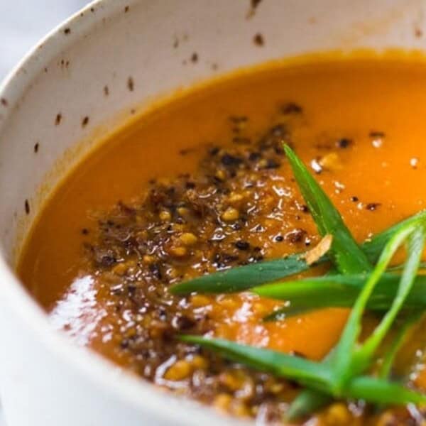 A close-up of a bowl of carrot soup, garnished with chopped scallions and ground black pepper. The soup is a vibrant orange color, and the bowl sits on a light surface.