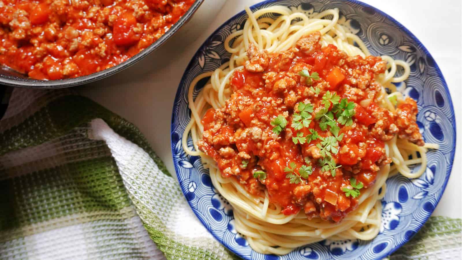 A blue plate of spaghetti topped with a rich, chunky tomato and meat sauce, garnished with fresh parsley. An adjacent dish with more sauce and a green and white striped cloth can be seen beside it.