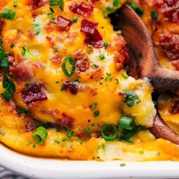 A casserole dish filled with cheesy mashed potatoes topped with crispy bacon bits and chopped green onions. A wooden spoon is scooping some out, revealing the creamy texture beneath the golden-brown crust.