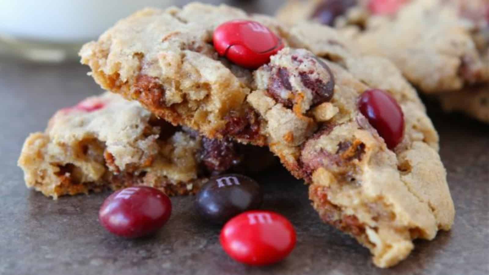 Close-up of a chocolate chip cookie broken in half, with red and brown candy-coated chocolates scattered on top and around it, placed on a dark surface.