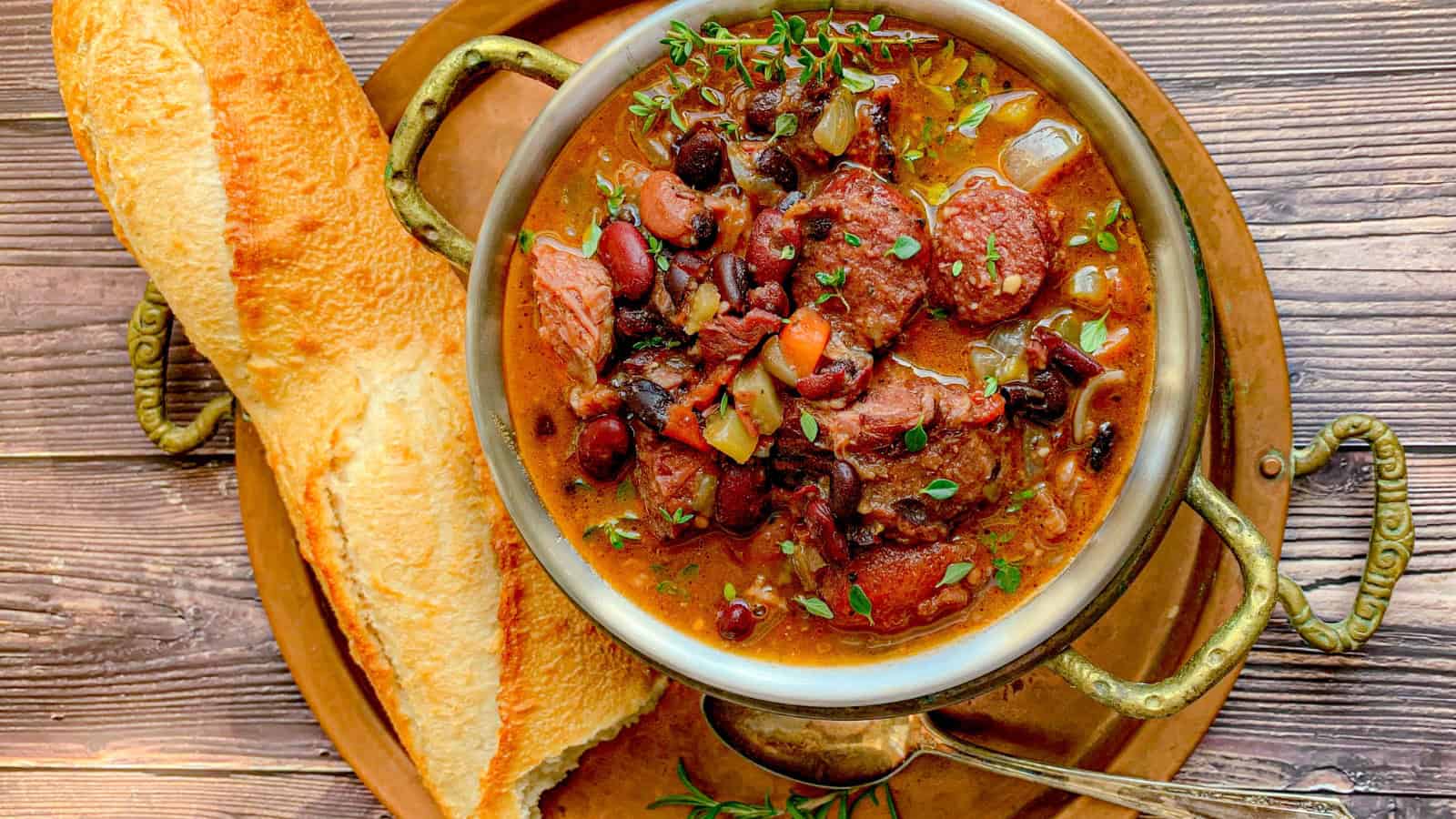 A bowl of hearty stew with sliced sausage, beans, and vegetables, garnished with herbs, sits on a copper plate. A slice of crusty baguette rests beside it. A spoon lies on the plate, ready to serve. The wooden table surface adds a rustic touch.