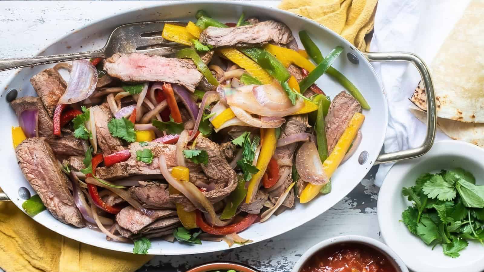 A mixed stir-fry of sliced beef, yellow and green bell peppers, red onion, and cilantro in a white serving dish with handles. A metal fork rests on top. On the side are fresh cilantro leaves and a small bowl of salsa.