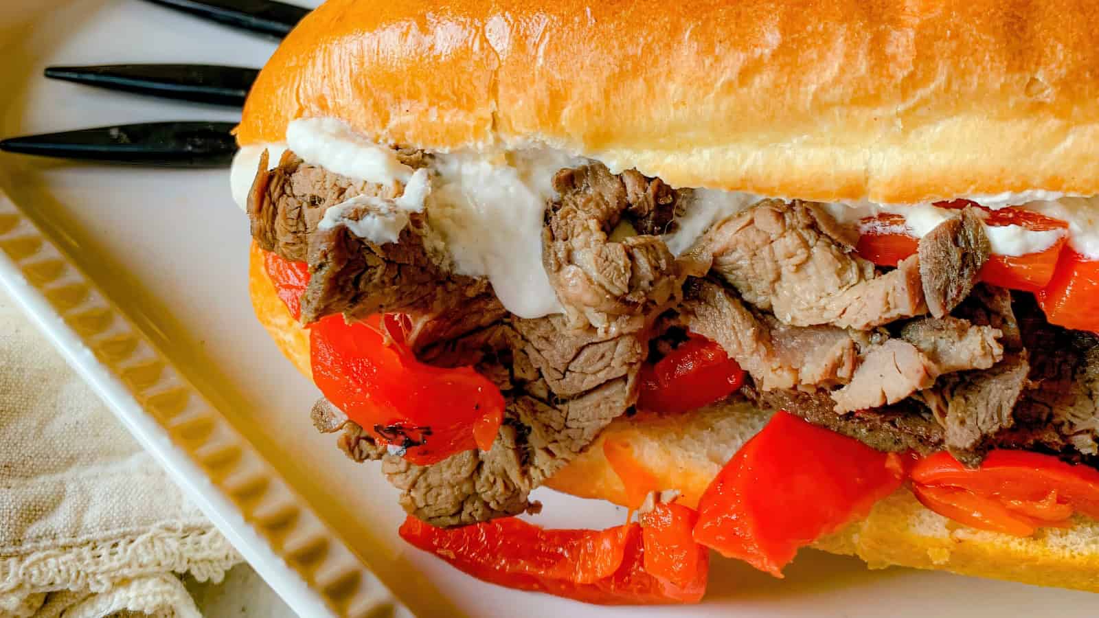 Close-up of a sandwich filled with sliced roast beef, red peppers, and a dollop of creamy sauce on a soft bun. The sandwich is on a white plate with black utensils in the background.
