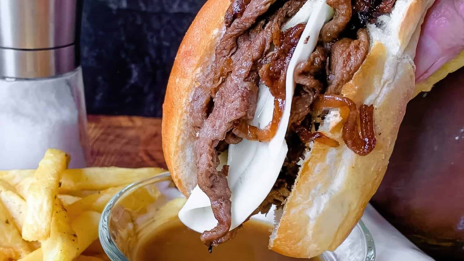 A close-up of a sandwich filled with slices of roast beef, caramelized onions, and melted cheese on a bun. It’s held above a bowl of gravy, with a side of golden fries on a wooden surface.
