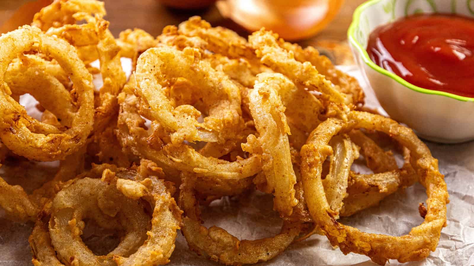 A close-up of crispy, golden-brown onion rings on parchment paper with a small bowl of red ketchup on the side. The onion rings are stacked irregularly and appear freshly fried.