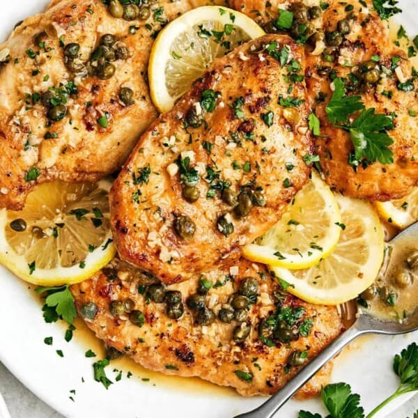 Plate of chicken piccata garnished with lemon slices, capers, and parsley. A spoon is placed on the plate, next to a fork and knife. Striped napkin on the side.