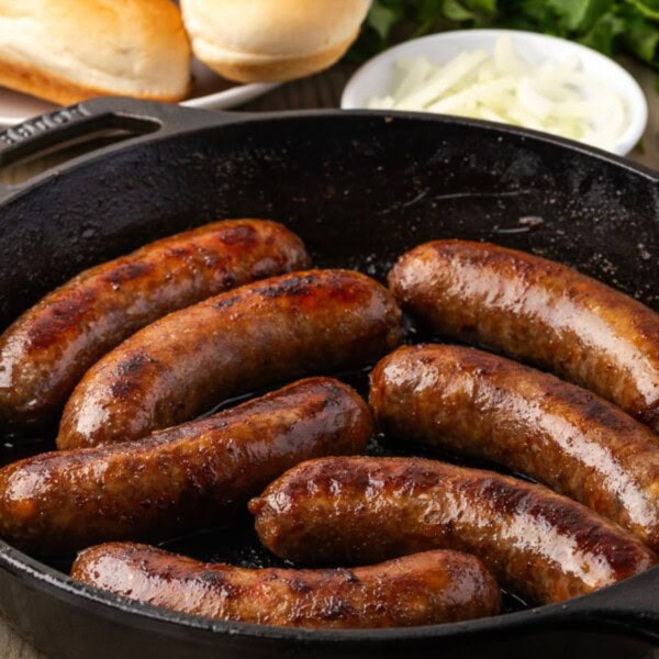 Seven browned sausages cooking in a black cast iron skillet, with mustard, bread, onions, and fresh herbs on a wooden table.