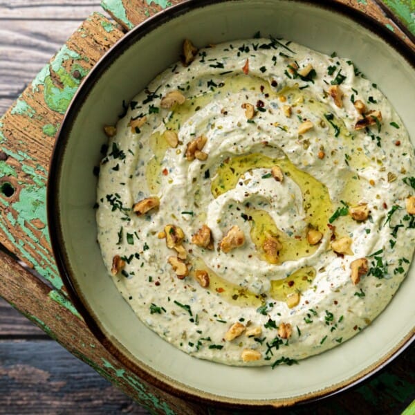 A bowl of creamy dip topped with chopped herbs, olive oil, and crushed nuts rests on a rustic green wooden chair.