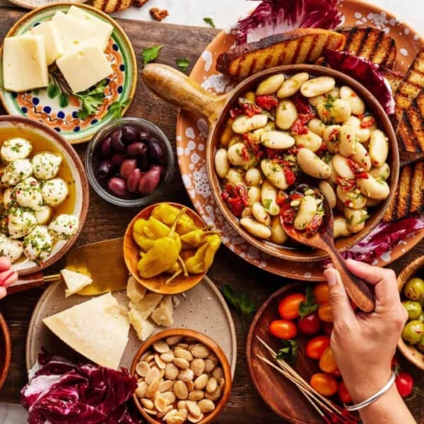 A vibrant spread of Mediterranean appetizers, including cheeses, olives, pickled vegetables, marinated beans, cherry tomatoes, and grilled bread. Two hands are serving food from the plates. Two glasses of red wine accompany the meal.