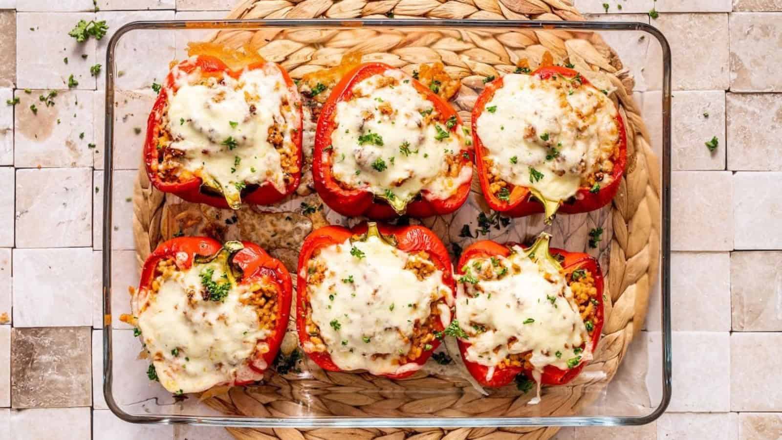 A baking dish filled with six red bell peppers, halved and stuffed with a rice and meat mixture, topped with melted cheese and garnished with parsley. The dish is placed on a woven mat on a tiled surface.