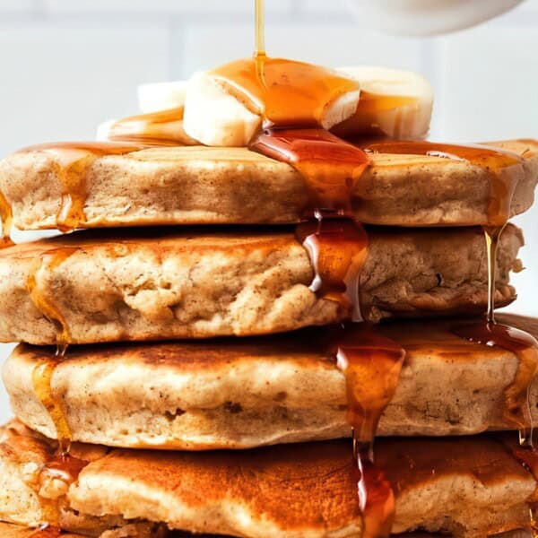 A stack of pancakes topped with banana slices and being drizzled with syrup. The pancakes are golden brown and the syrup is flowing down the sides. A blurred background features a white mug and a tiled wall.