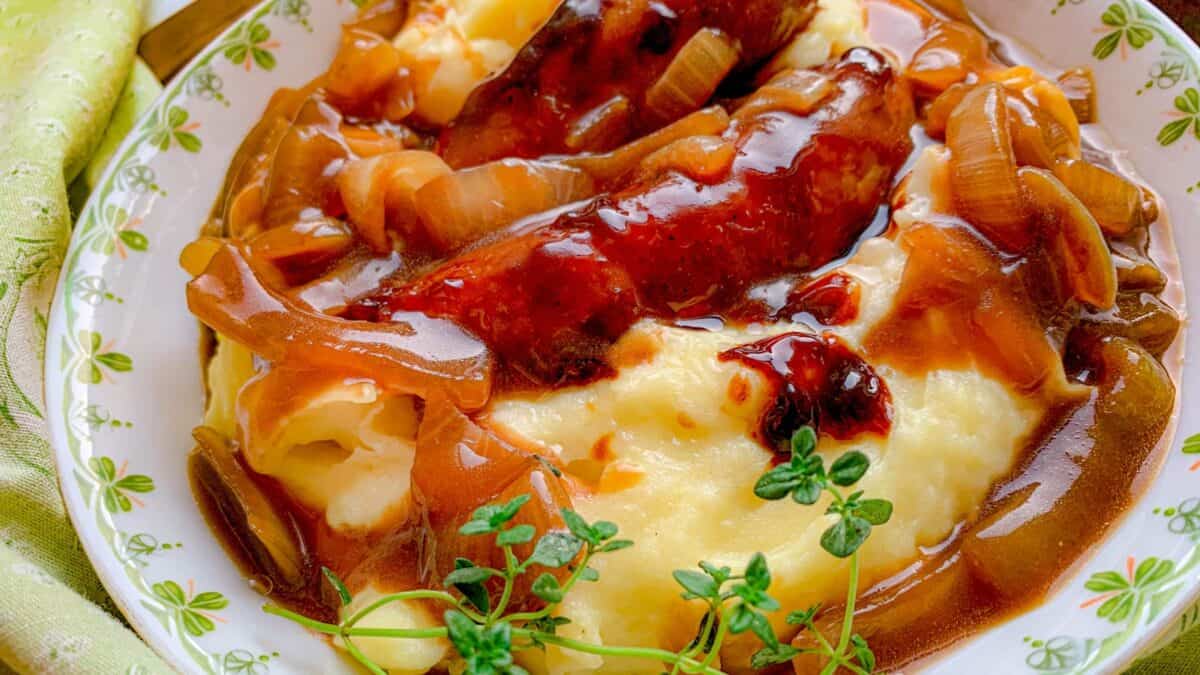 A plate of mashed potatoes topped with sausages, caramelized onions, brown gravy, and a sprig of fresh thyme.