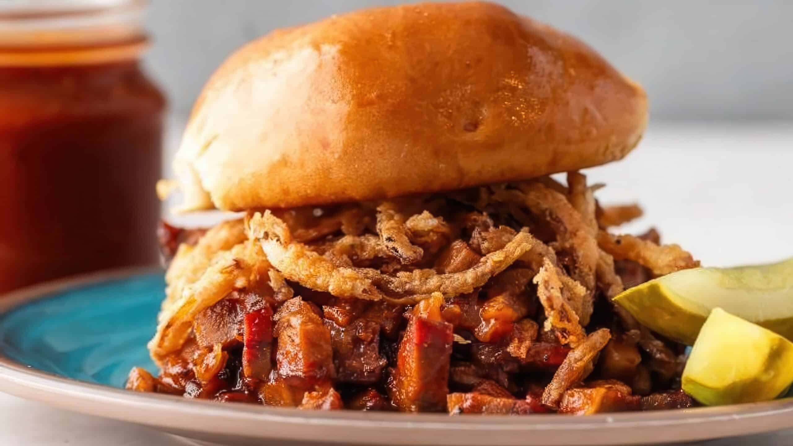 A BBQ sandwich with a glossy bun, filled with chopped barbecued meat and crispy fried onions, served on a teal plate with pickle slices on the side. A jar of sauce is in the background.