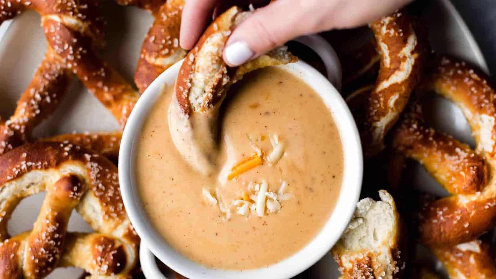 A hand dips a soft pretzel into a bowl of cheese sauce, surrounded by more pretzels on a plate. The cheese sauce has small shreds of cheese on top.