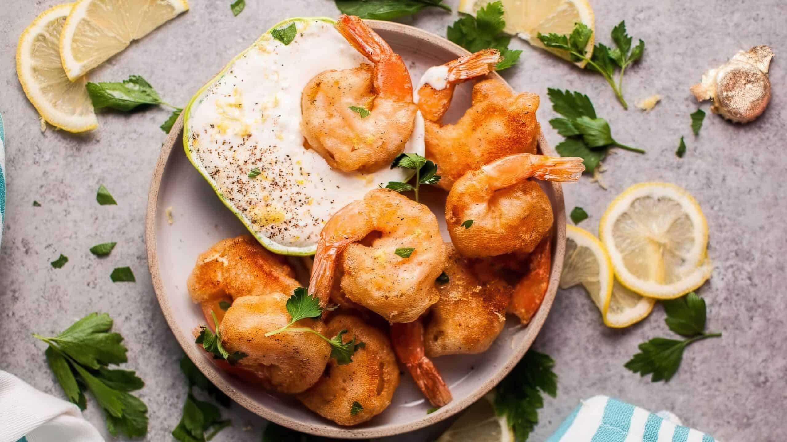 A plate of crispy fried shrimp garnished with fresh parsley, accompanied by a dish of creamy dipping sauce. Lemon slices and parsley leaves are scattered around on a textured gray surface.