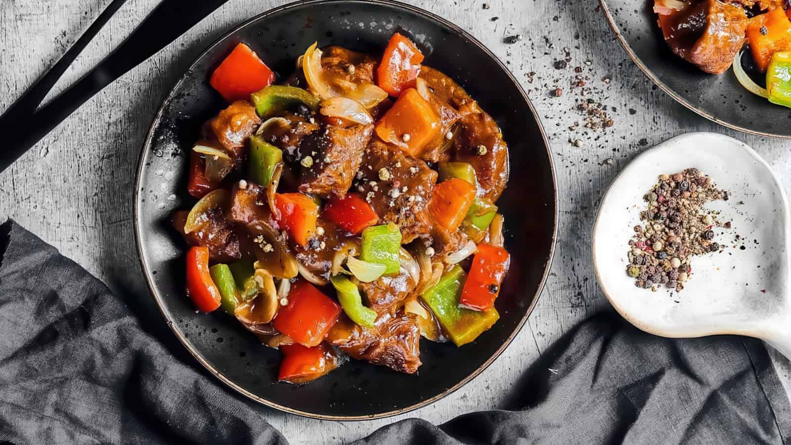 A black plate with stir-fried chunks of beef, red and green bell peppers, and onions, all coated in a savory sauce. Black chopsticks are placed beside the plate. A small dish with mixed peppercorns is on the right, and a dark napkin is nearby.