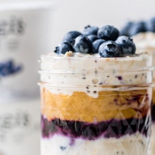 A close-up of a layered parfait in a jar, featuring yogurt, peanut butter, blueberry sauce, chia seeds, and topped with fresh blueberries. Blurred yogurt containers are in the background.