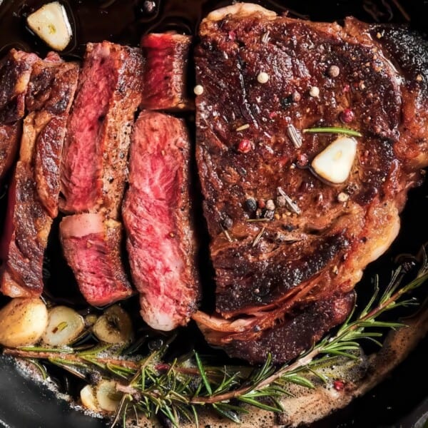 A perfectly cooked steak sliced in a cast iron skillet, topped with garlic cloves and rosemary. The juicy, seared meat displays a medium-rare center, surrounded by a crispy crust, with a cloth napkin nearby.