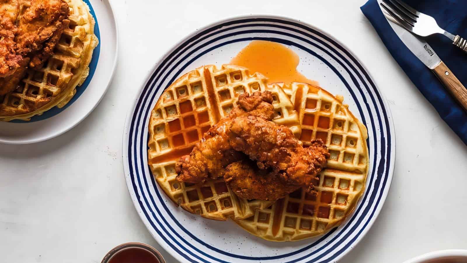 A plate with two golden waffles topped with crispy fried chicken and drizzled with syrup. A fork and knife sit on a blue napkin beside the plate, and part of another plate with waffles is visible in the background.