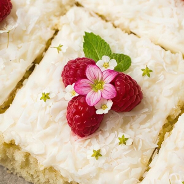 Sheet cake slices topped with white frosting, shredded coconut, raspberries, pink flowers, and mint leaves.