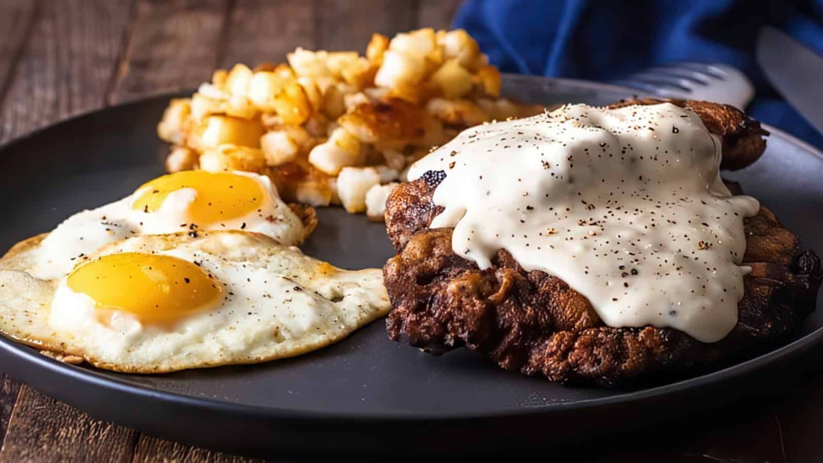 A plate with two sunny-side-up eggs, crispy hash browns, and a piece of fried meat topped with creamy gravy. The dish is served on a dark round plate on a wooden table.