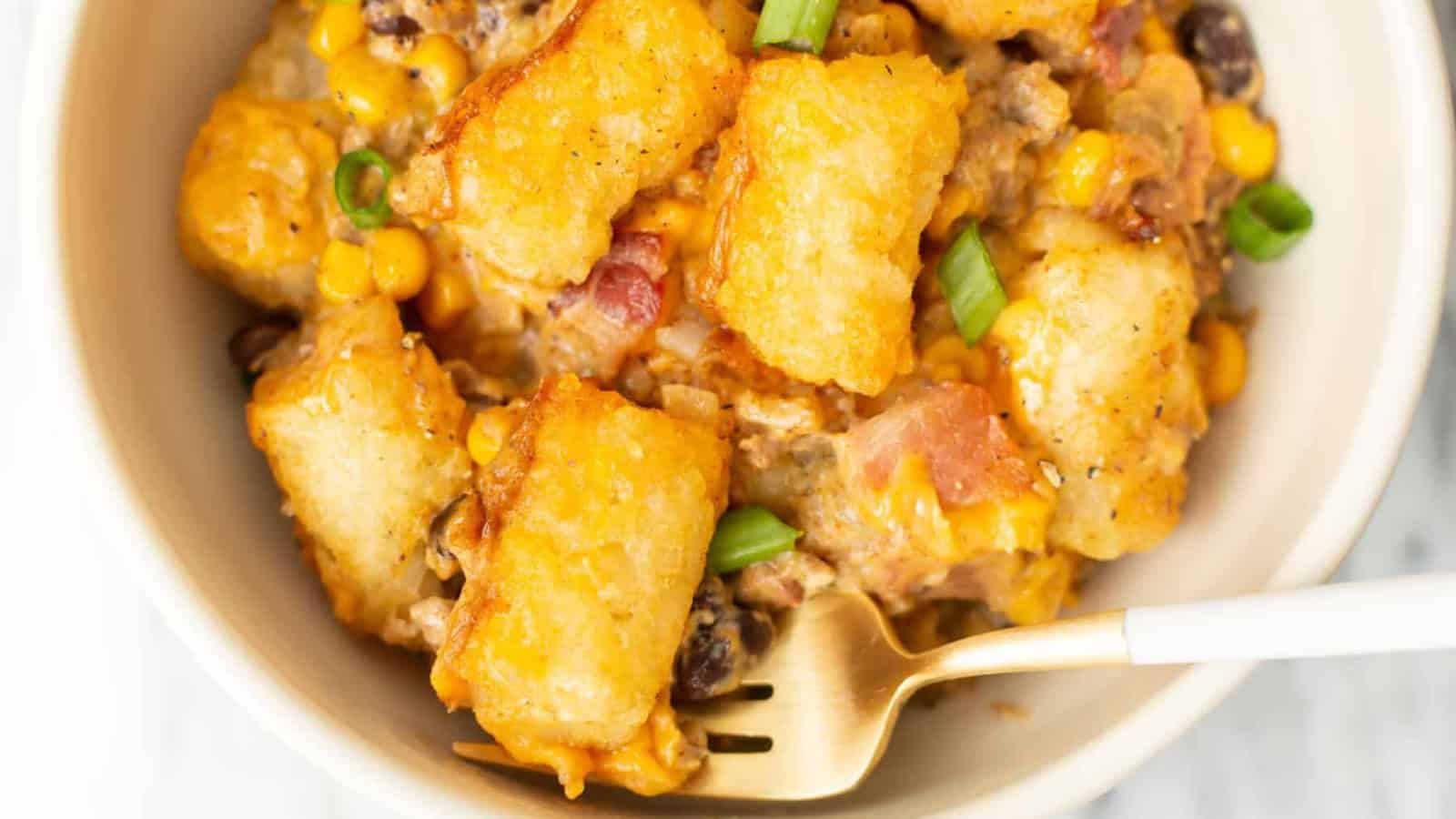 A bowl of tater tot casserole with golden-brown tots on top, mixed with corn, black beans, and diced red tomatoes. Garnished with chopped green onions, a fork is placed on the right side of the bowl.