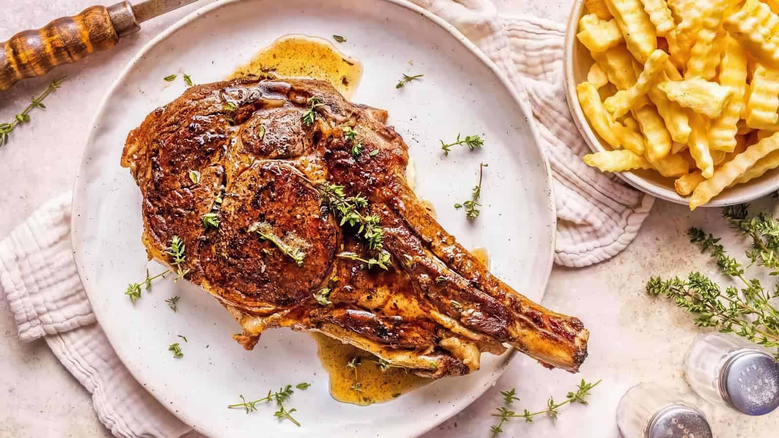 A seared ribeye steak garnished with herbs sits on a white plate with a small pool of sauce. To the side, there is a bowl of crinkle-cut French fries. The table is adorned with a white cloth and sprigs of fresh herbs.