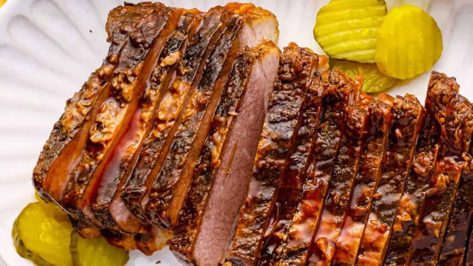 Sliced brisket with a caramelized crust is arranged on a white plate next to sliced pickles.