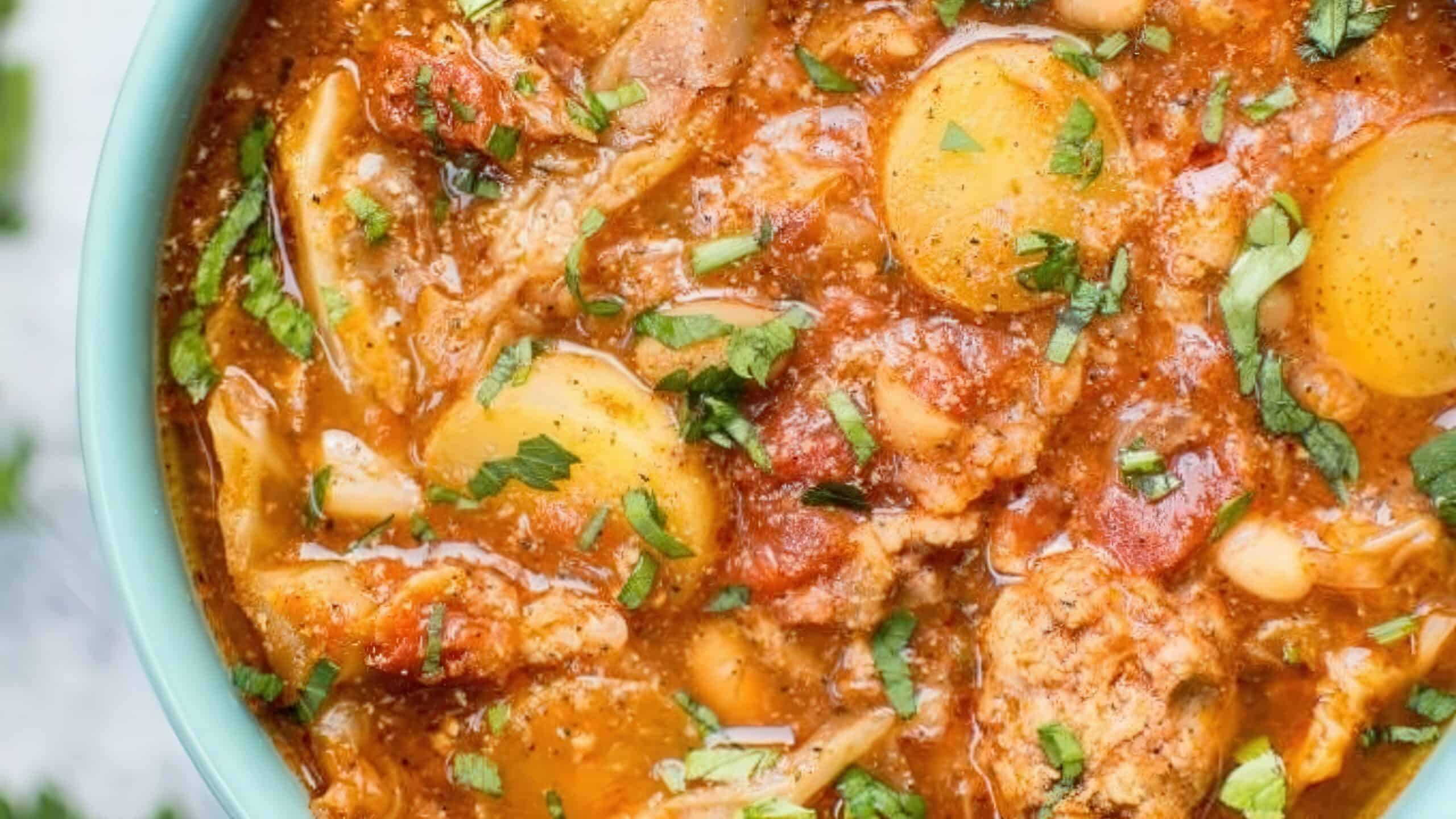 A close-up of a vibrant stew in a bowl, featuring yellow potatoes, shredded cabbage, tomatoes, and white beans. The dish is garnished with fresh chopped herbs, creating a colorful and appetizing appearance.