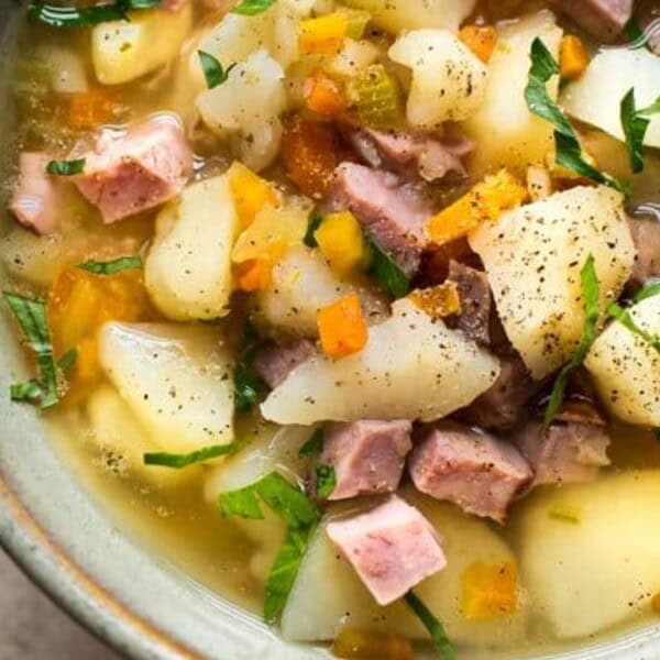 A bowl of hearty stew filled with diced potatoes, chunks of meat, and sliced vegetables, garnished with fresh herbs and pepper, presented in a rustic ceramic bowl.