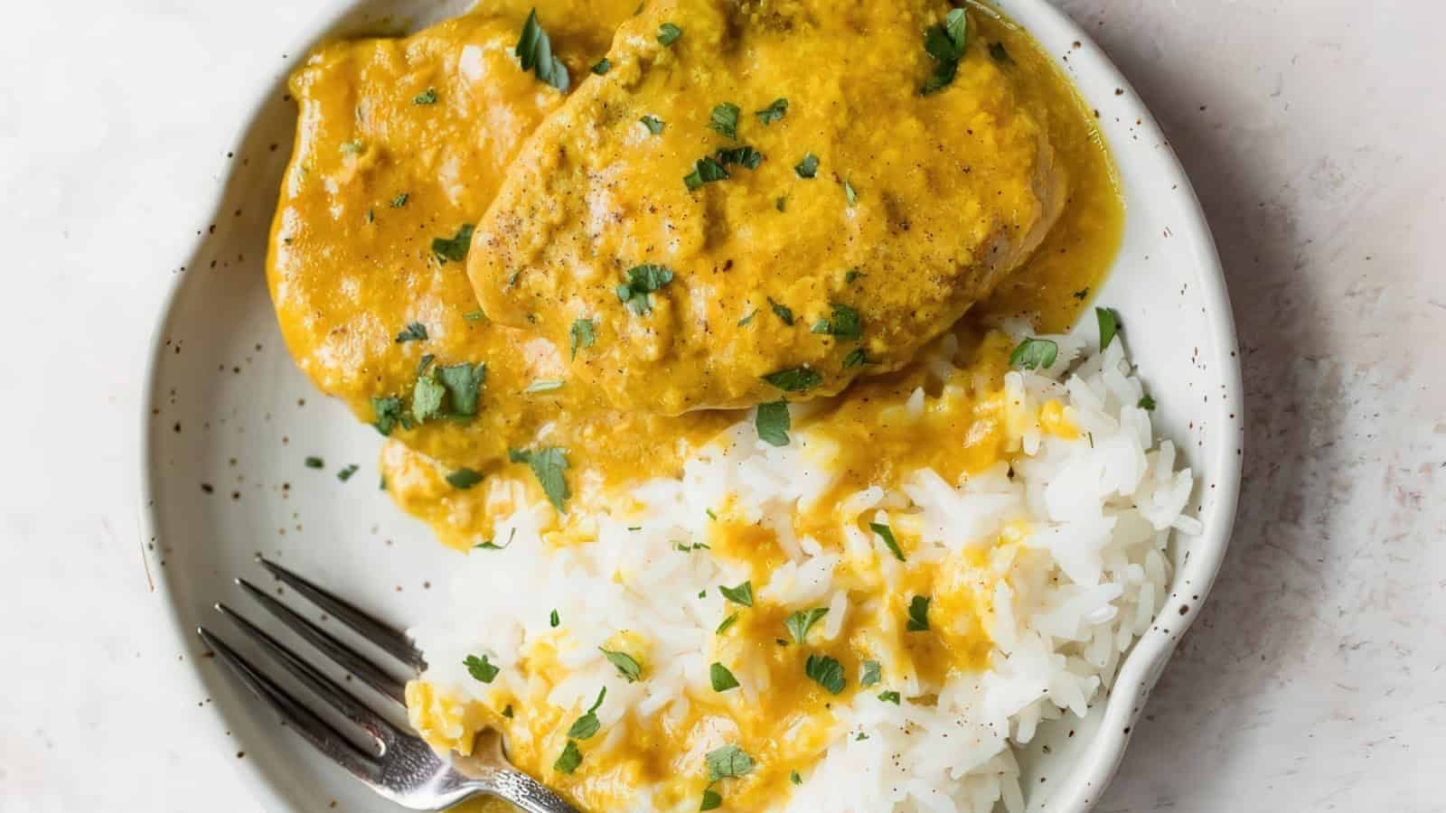 A white plate with rice and chicken covered in a creamy yellow curry sauce, garnished with chopped parsley. A fork rests on the side of the plate.