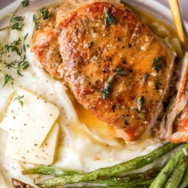 A plate featuring a thick piece of grilled chicken garnished with herbs, served with a side of creamy mashed potatoes topped with butter, and sautéed green beans. The dish is presented on a light-colored surface.