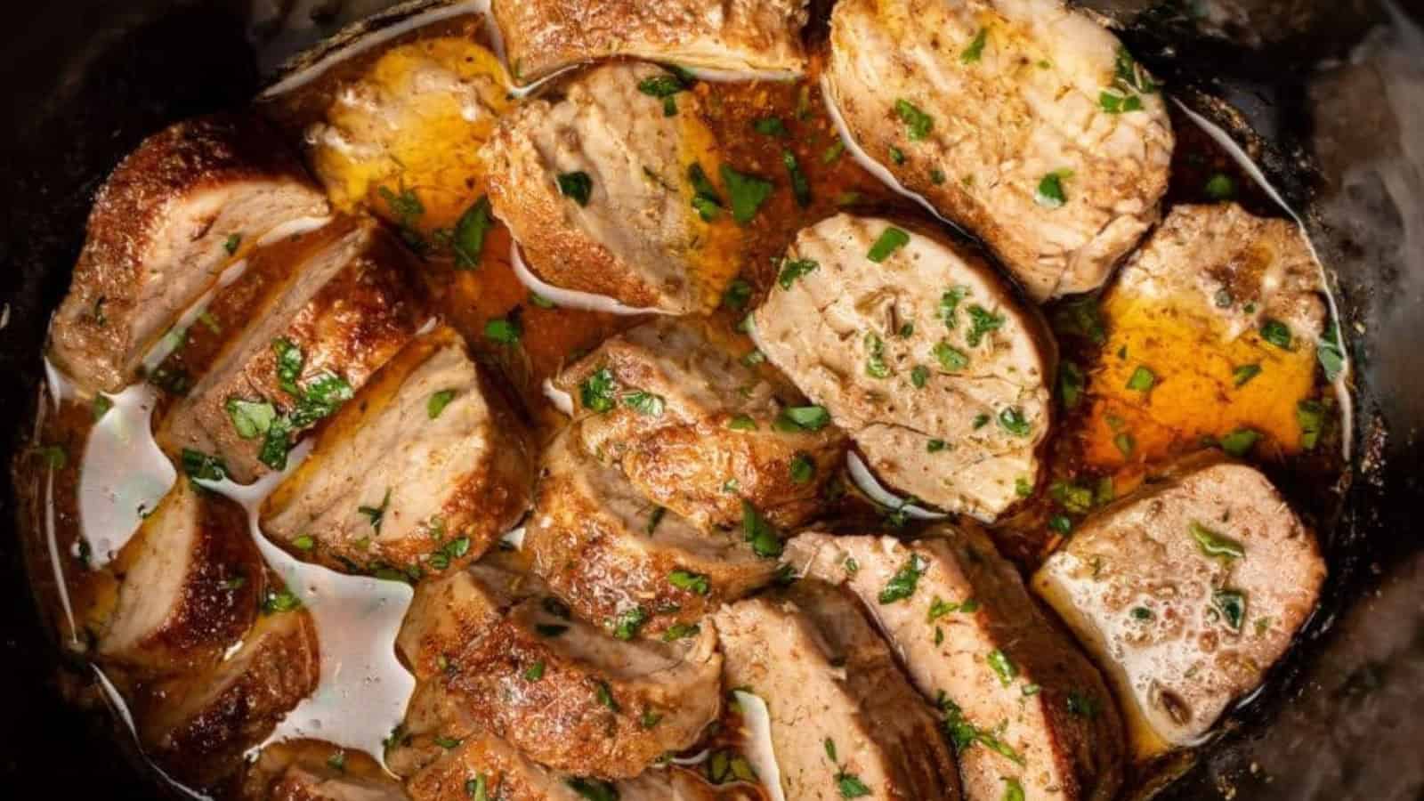 Sliced cooked pork tenderloin in a rich, brown sauce, garnished with fresh herbs. The meat is juicy and appears well-seasoned, surrounded by a flavorful liquid.