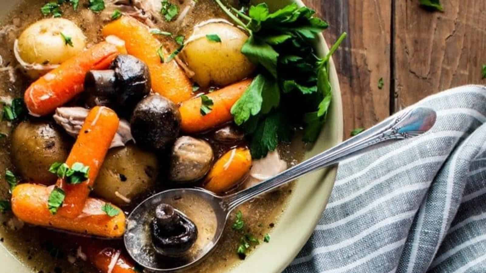 A bowl of hearty stew filled with whole baby carrots, potatoes, mushrooms, and tender meat in a rich broth garnished with chopped parsley. A striped napkin and spoon rest beside the bowl on a rustic wooden table.