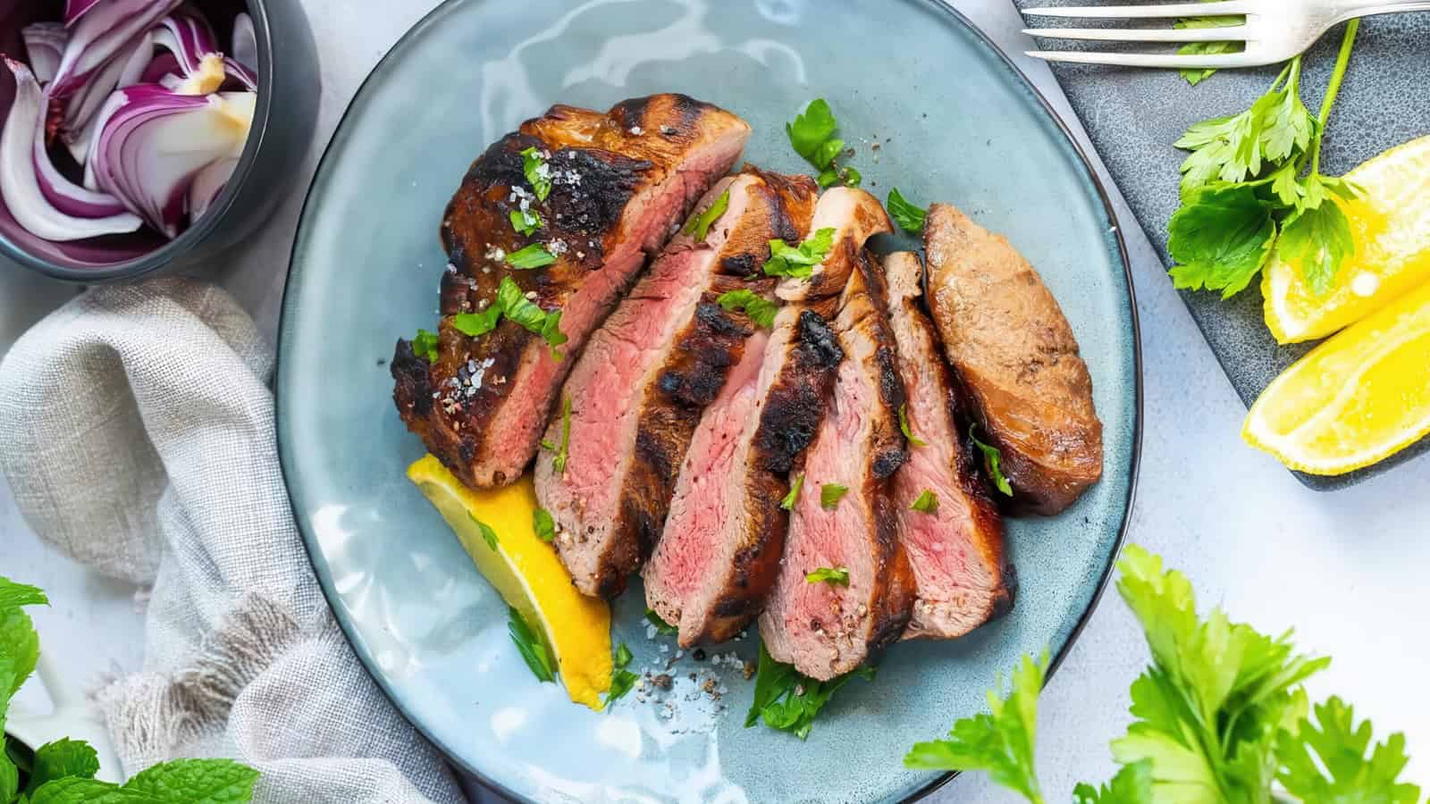 A plate with sliced medium-rare grilled steak garnished with fresh herbs. Lemon wedges are on the side. A bowl of sliced red onions and a fork are visible nearby. The setting includes a light gray napkin and green leaves scattered around.