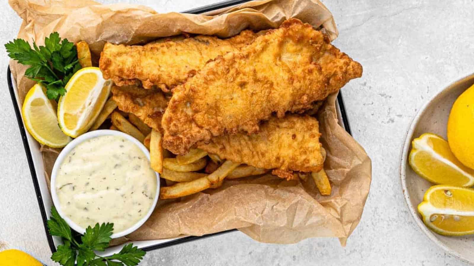 A tray of crispy golden fish and chips served with lemon wedges and tartar sauce, garnished with parsley, on a parchment-lined container. A plate of lemons is nearby on a light gray surface.