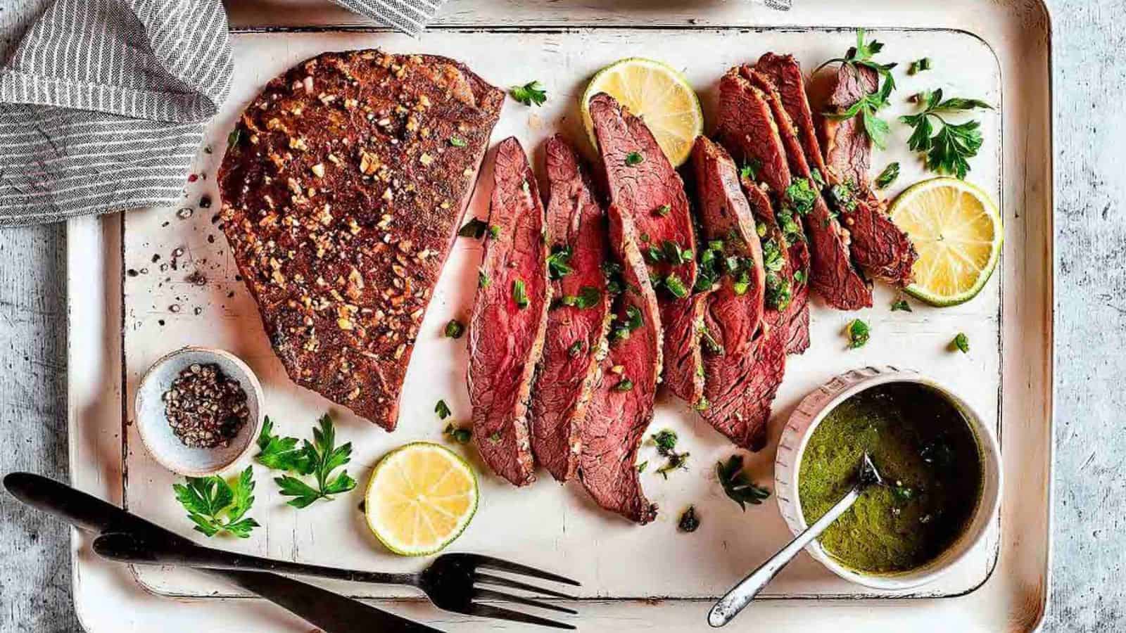 A platter with sliced grilled steak garnished with herbs, surrounded by lemon slices and parsley. A small bowl of chimichurri sauce and a dish of pepper are on the side. A fork and a striped cloth are nearby.