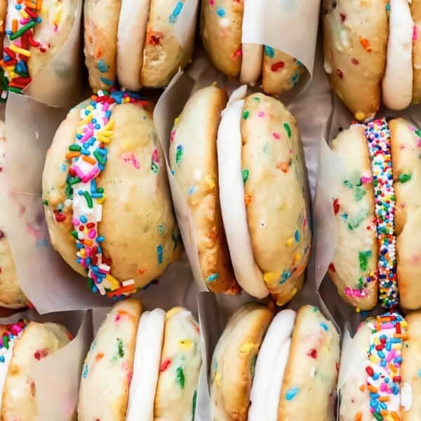 A close-up of colorful whoopie pies. Each pie consists of two round cookies with a white cream filling. The cookies are speckled with rainbow sprinkles, and additional sprinkles decorate the cream filling, creating a festive appearance.