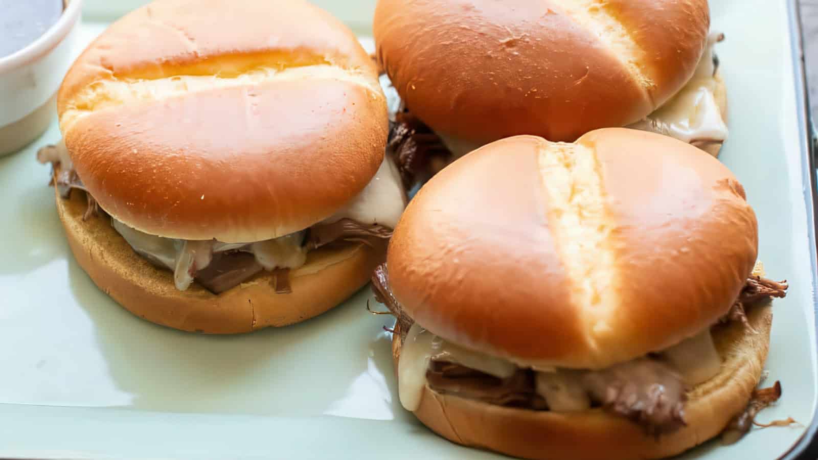Three beef sandwiches with melted cheese on soft buns are placed on a light-colored plate. A small container of dipping sauce is partially visible in the top left corner.